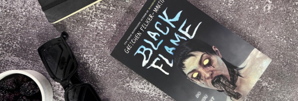 A paperback copy of the book Black Flame by Gretchen Felker-Martin sitting on a distressed cement background. A notebook, pen, container of blackberries, and black sunglasses are laid out near it.
